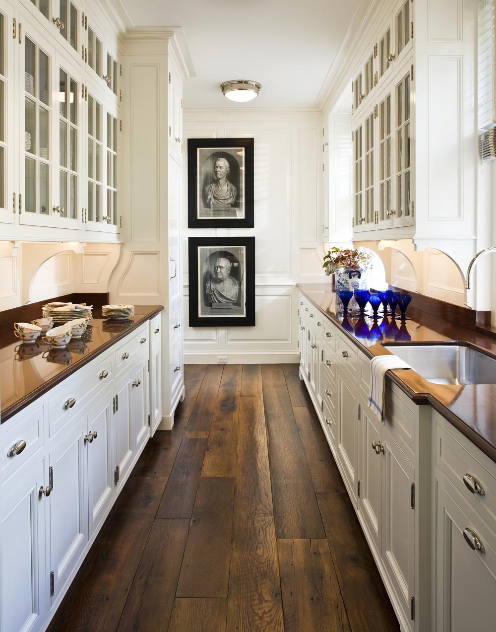 Bright white kitchen with glass-front cabinets, a modern faucet over a sink, a bowl of oranges, gray plates, cutlery, glasses, a black bottle, a metal jar, and a vase with white flowers on a marble countertop.