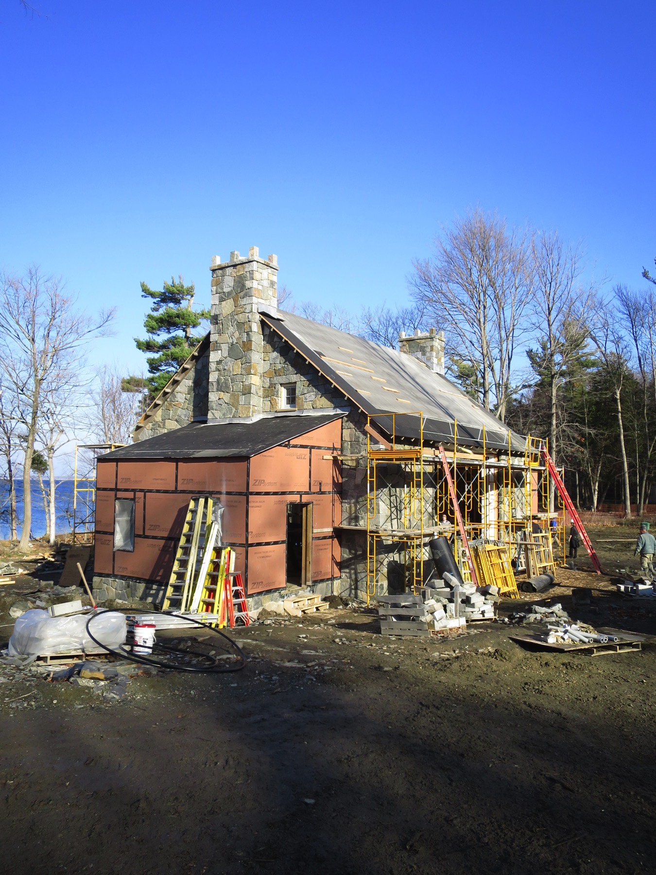A small stone house with a gray roof stands among leafing trees. Bare soil and a mound of dirt surround the house. The sky is clear and light blue, and young spring foliage covers the trees in the background.
